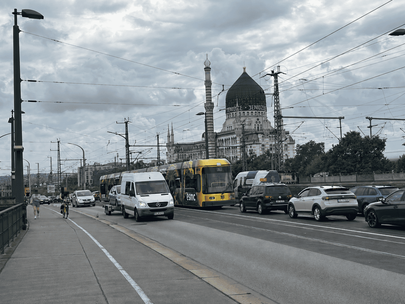 Eine viel befahrene Straße auf einer Brücke mit Autos und einer gelben Straßenbahn. Im Hintergrund steht das historische Gebäude Yenidze mit seiner markanten Kuppel unter einem bewölkten Himmel. Links befindet sich ein Gehweg.