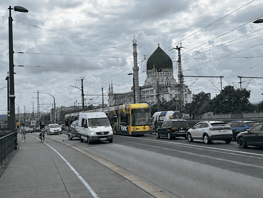 Eine viel befahrene Straße auf einer Brücke mit Autos und einer gelben Straßenbahn. Im Hintergrund steht das historische Gebäude Yenidze mit seiner markanten Kuppel unter einem bewölkten Himmel. Links befindet sich ein Gehweg.