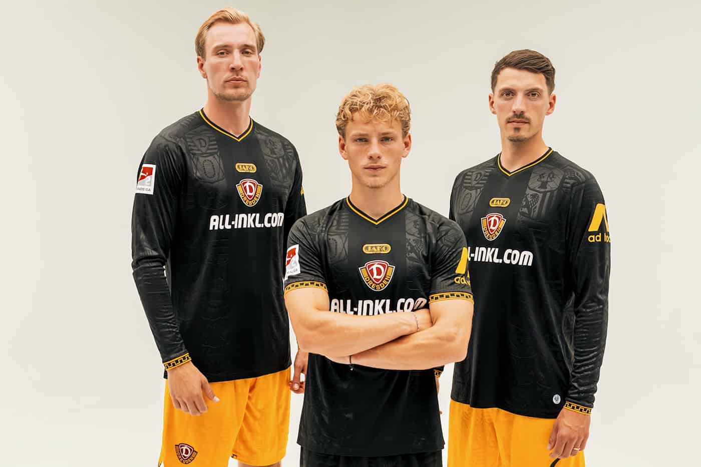 Drei Fußballspieler von Dynamo Dresden stehen nebeneinander vor weißem Hintergrund. Sie tragen das neue schwarze Auswärtstrikot mit gelben Bündchen und dem Vereinswappen in der Mitte.