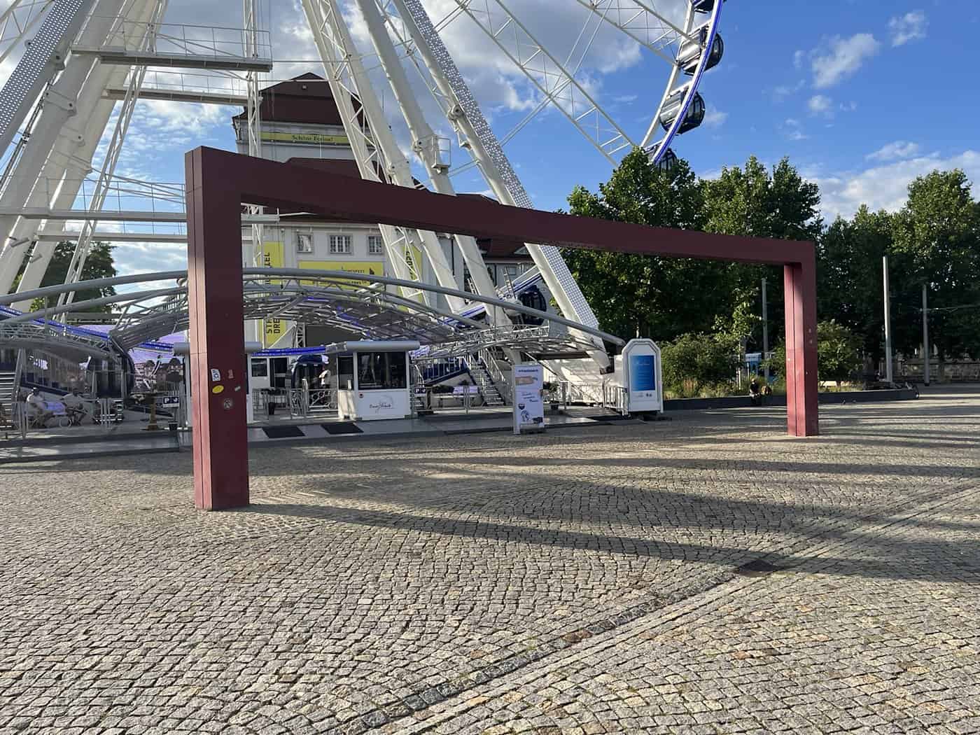 Ein großer, eckiger Rahmen aus rotem Metall steht auf einem gepflasterten Platz. Direkt dahinter ragt das weiße Gerüst eines riesigen Riesenrads in den leicht bewölkten blauen Himmel.