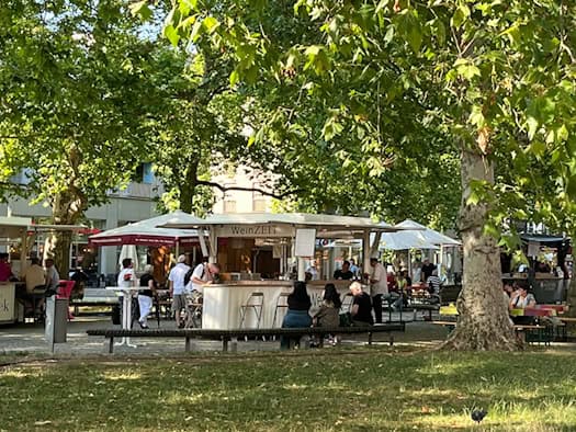Ein schattiger Festplatz unter großen, grünen Bäumen. Im Hintergrund stehen weiße Verkaufsbuden wie der Stand „WeinZEIT“ und farbige Biertische, an denen Menschen sitzen. Im sonnigen Gras im Vordergrund picken Tauben.
