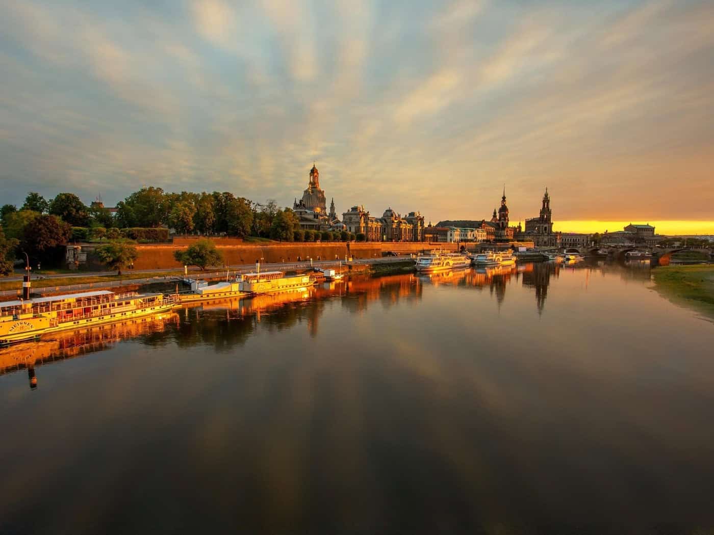 Die historische Altstadt von Dresden mit der Frauenkirche spiegelt sich bei goldenem Sonnenuntergang im ruhigen Wasser der Elbe, an deren Ufer mehrere Schiffe anlegen.