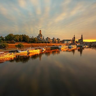 Die historische Altstadt von Dresden mit der Frauenkirche spiegelt sich bei goldenem Sonnenuntergang im ruhigen Wasser der Elbe, an deren Ufer mehrere Schiffe anlegen.