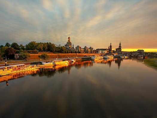 Die historische Altstadt von Dresden mit der Frauenkirche spiegelt sich bei goldenem Sonnenuntergang im ruhigen Wasser der Elbe, an deren Ufer mehrere Schiffe anlegen.