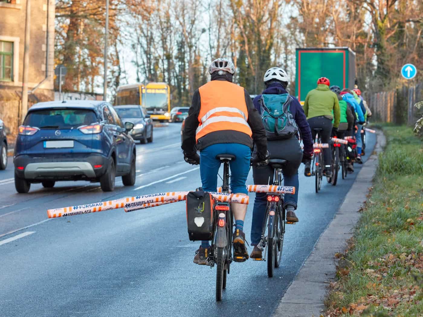 Radfahrer fahren in einer Reihe auf einer Straße. An ihren Gepäckträgern sind orangefarbene Poolnudeln quer befestigt, die seitlich in den Fahrbahnraum ragen, um den geforderten Überholabstand zu Autos sichtbar zu machen.