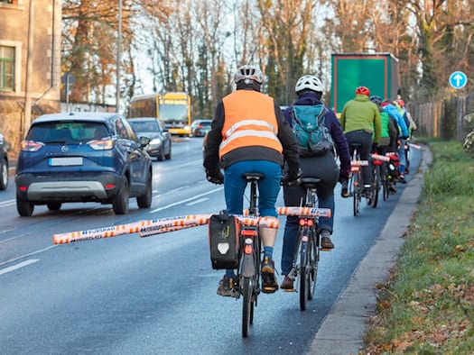 Radfahrer fahren in einer Reihe auf einer Straße. An ihren Gepäckträgern sind orangefarbene Poolnudeln quer befestigt, die seitlich in den Fahrbahnraum ragen, um den geforderten Überholabstand zu Autos sichtbar zu machen.