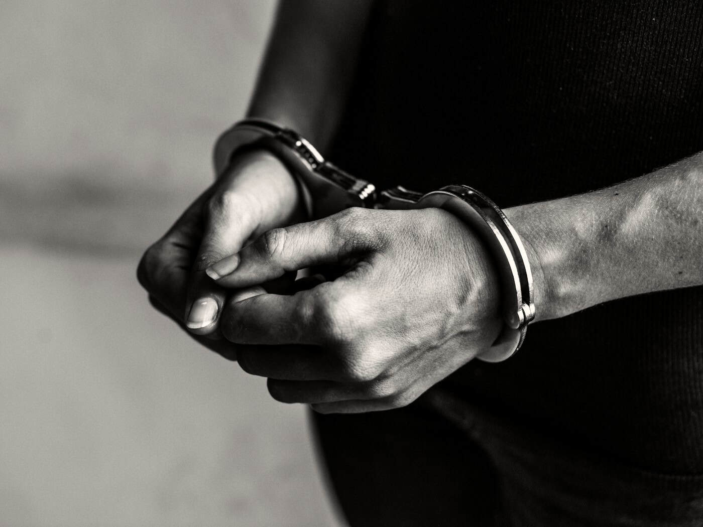 Schwarz-Weiß-Nahaufnahme von zwei menschlichen Händen, die vor dem Oberkörper gefesselt in metallenen Handschellen liegen. Der Hintergrund ist schlicht und dunkel gehalten.