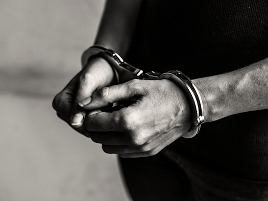 Schwarz-Weiß-Nahaufnahme von zwei menschlichen Händen, die vor dem Oberkörper gefesselt in metallenen Handschellen liegen. Der Hintergrund ist schlicht und dunkel gehalten.