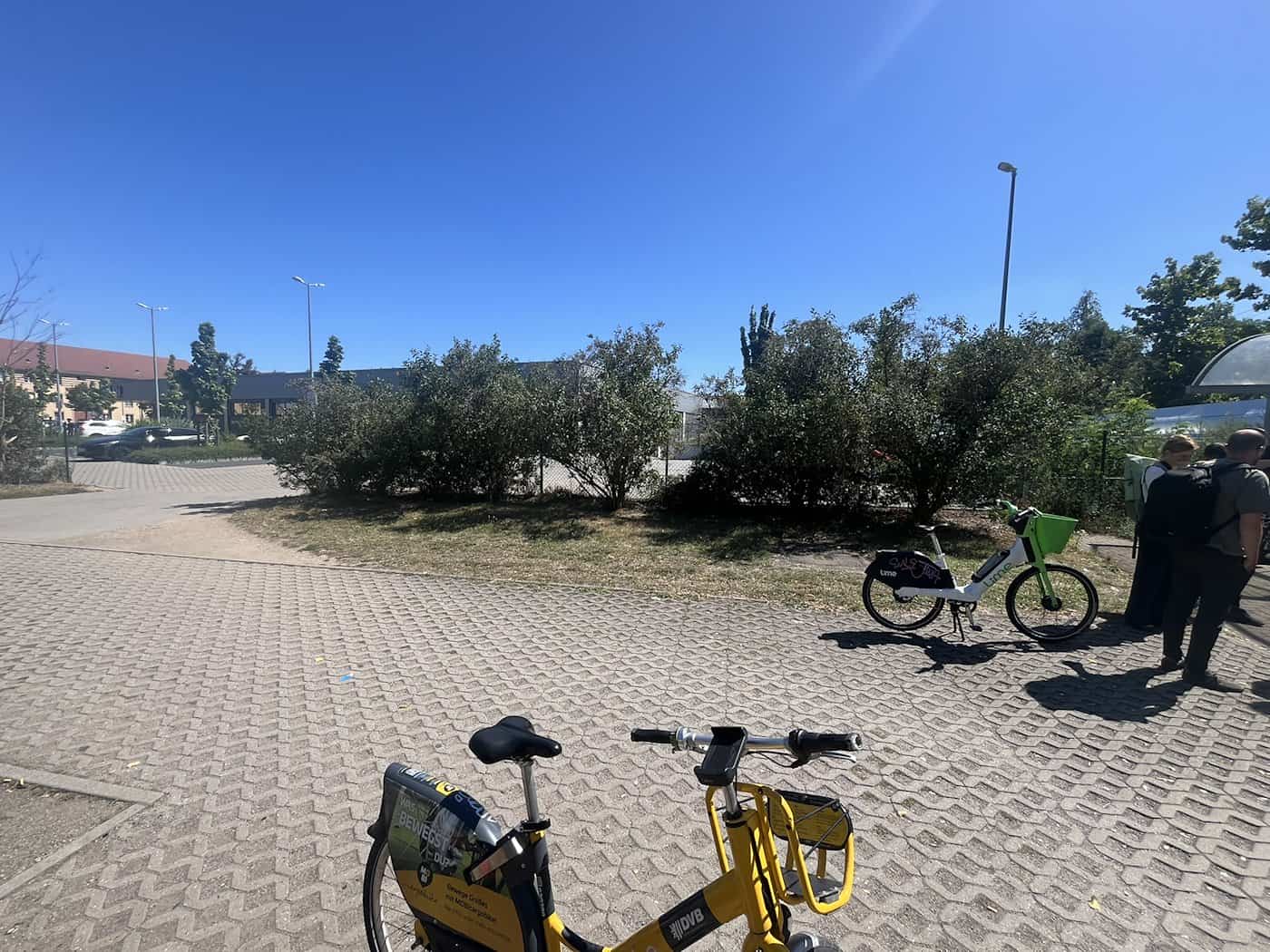 Gepflastertes Areal am S-Bahnhof Dresden-Dobritz bei strahlendem Sonnenschein. Im Vordergrund parkt ein gelbes Leihfahrrad, im Hintergrund sind weitere Räder, Büsche und Passanten zu sehen.