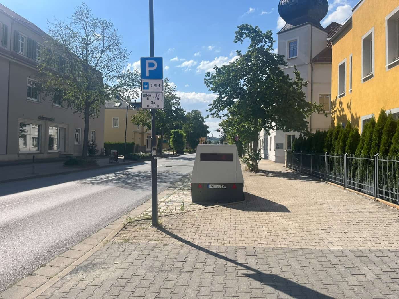 Ein grauer Blitzeranhänger mit dem Kennzeichen HWI VE 159 steht auf dem Gehweg der Dresdner Straße in Dresden. Rechts befindet sich ein gelbes Gebäude mit einem kleinen Turm, links verläuft die Fahrbahn an einem blauen Parkplatzschild vorbei.