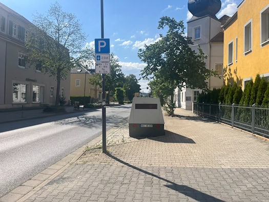 Ein grauer Blitzeranhänger mit dem Kennzeichen HWI VE 159 steht auf dem Gehweg der Dresdner Straße in Dresden. Rechts befindet sich ein gelbes Gebäude mit einem kleinen Turm, links verläuft die Fahrbahn an einem blauen Parkplatzschild vorbei.
