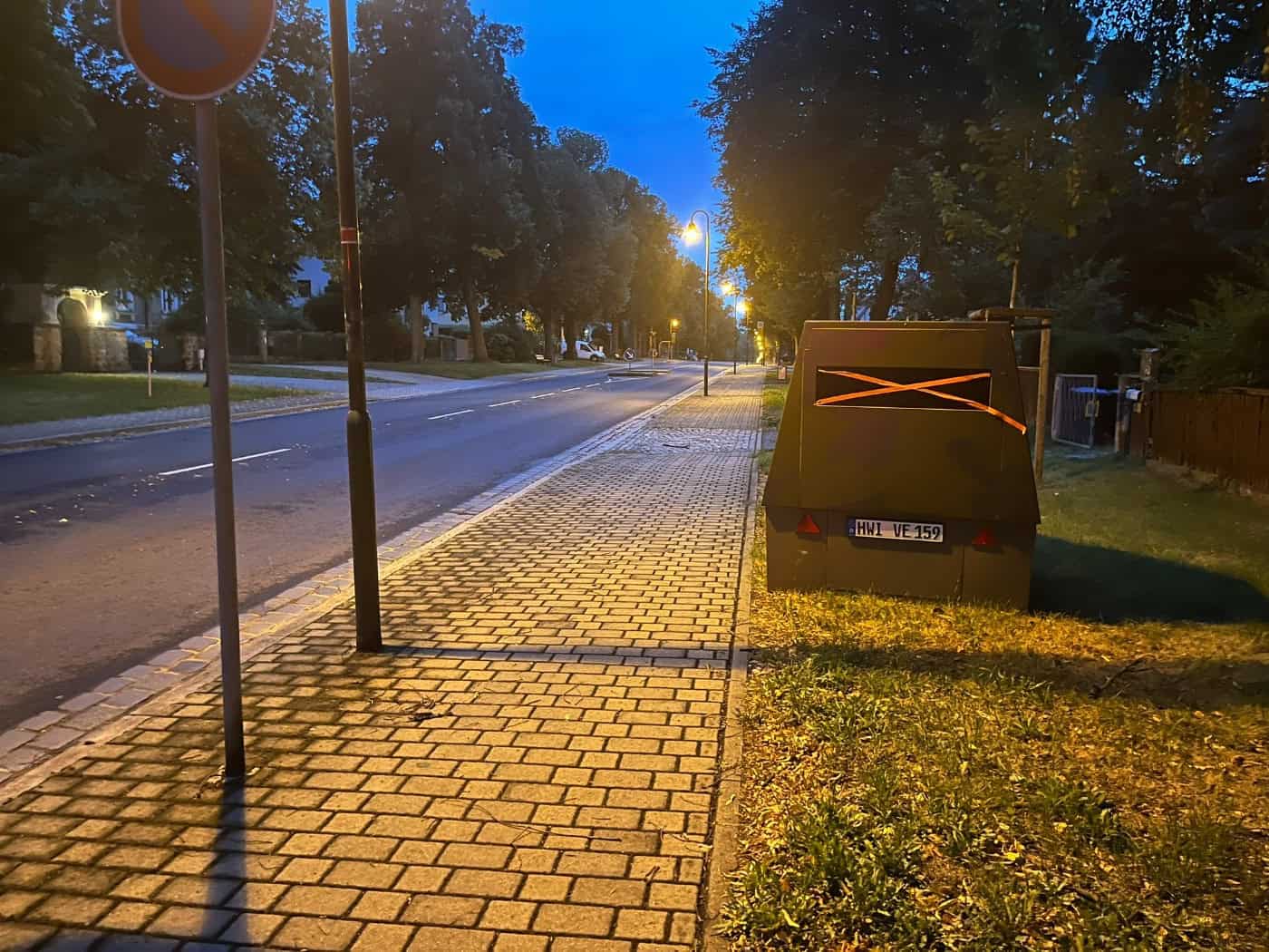 Ein olivgrüner Blitzeranhänger mit dem Kennzeichen HWI-VE 159 steht in der Dämmerung am Gehwegrand einer gepflasterten Straße. Über dem dunklen Kameraschlitz ist ein auffälliges orangefarbenes Kreuz aus Klebeband angebracht.