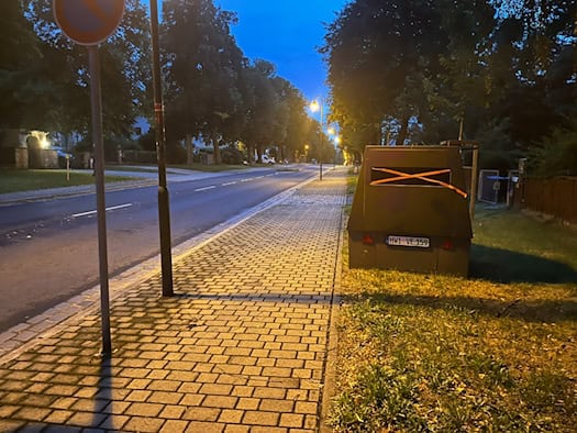 Ein olivgrüner Blitzeranhänger mit dem Kennzeichen HWI-VE 159 steht in der Dämmerung am Gehwegrand einer gepflasterten Straße. Über dem dunklen Kameraschlitz ist ein auffälliges orangefarbenes Kreuz aus Klebeband angebracht.