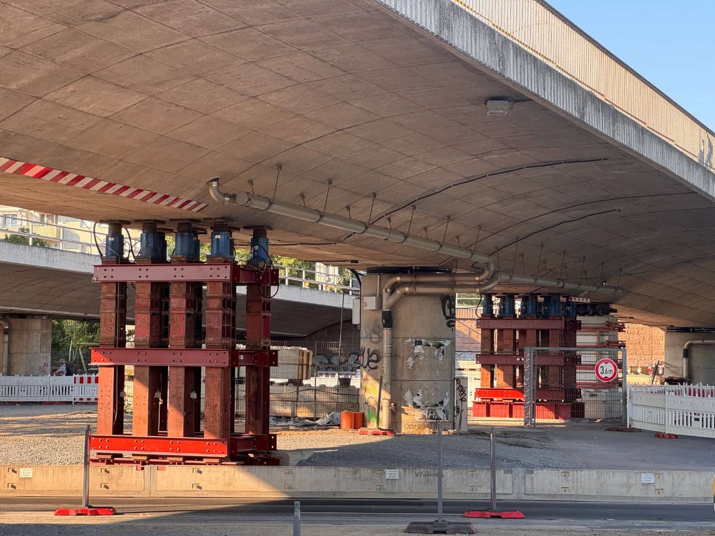 Unteransicht einer Betonbrücke, die von massiven, rot lackierten Stahlgerüsten und blauen Hydraulikhebern gestützt wird. Im Hintergrund sind Betonpfeiler, Baustellenabsperrungen und Schilder zu sehen.