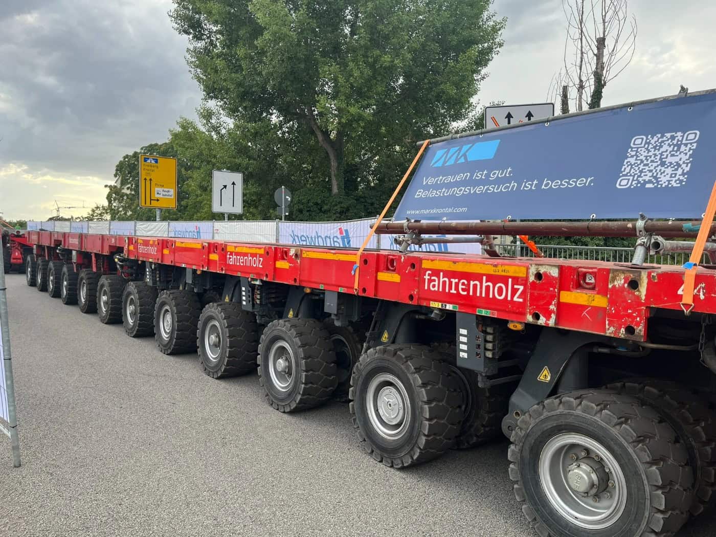 Seitenansicht eines roten Mehrachs-Schwerlasttransporters der Firma Fahrenholz auf der Brücke Budapester Straße. Auf dem Tieflader ist ein blaues Banner der Firma Marx Krontal mit der Aufschrift 'Vertrauen ist gut. Belastungsversuch ist besser' montiert.