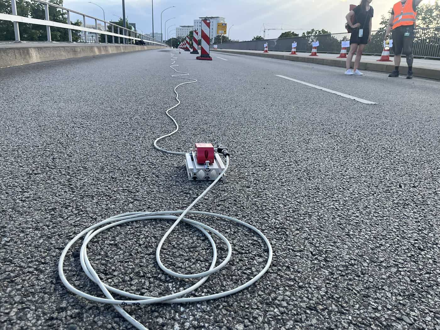 Nahaufnahme der asphaltierten Fahrbahn der Brücke Budapester Straße in Dresden. Ein roter Senkungssensor ist auf einer Metallhalterung befestigt, von dem ein weißes Kabel über die Straße verläuft. Im Hintergrund stehen Personen in Warnwesten.