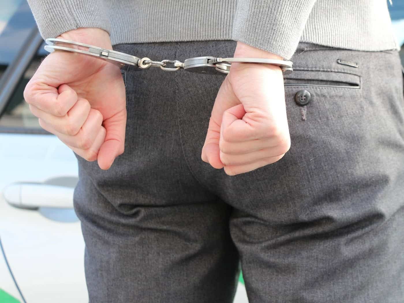 Symbolfoto: Rückansicht einer Person in grauer Hose und grauem Oberteil, deren Hände hinter dem Rücken auf Hüfthöhe in metallenen Handschellen gefesselt sind. Im Hintergrund ist die Lackierung eines Polizeifahrzeugs zu sehen.