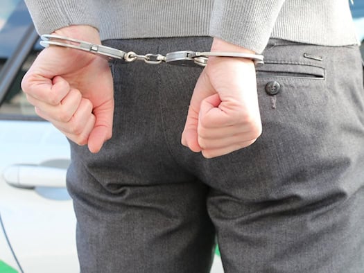 Symbolfoto: Rückansicht einer Person in grauer Hose und grauem Oberteil, deren Hände hinter dem Rücken auf Hüfthöhe in metallenen Handschellen gefesselt sind. Im Hintergrund ist die Lackierung eines Polizeifahrzeugs zu sehen.