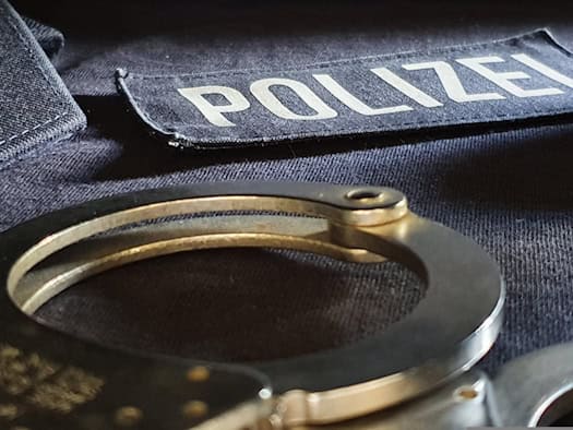 Nahaufnahme von Polizeiausrüstung: Im Vordergrund liegen metallische Handschellen auf dunklem Stoff. Dahinter ist ein Aufnäher mit der weißen Aufschrift „POLIZEI“ zu sehen. Im Hintergrund stapeln sich hellblaue Uniformhemden.