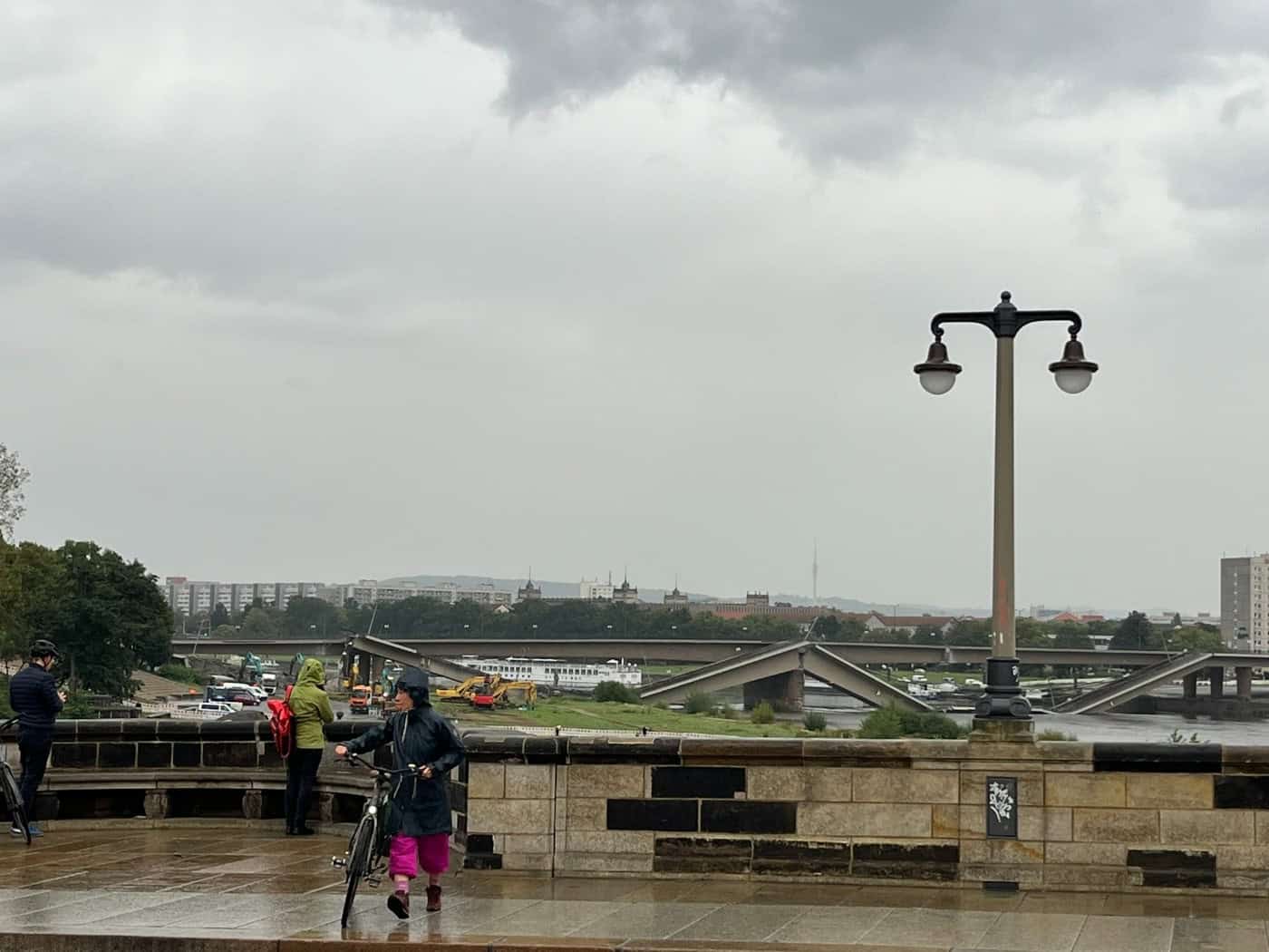 Passanten bei Regen mit Blick auf die eingestürzte Carolabrücke in Dresden. Eine Person schiebt ein Fahrrad über nasses Pflaster vor düsterem Himmel. Im Flussbett der Elbe liegen die abgebrochenen Betonteile der Brücke.
