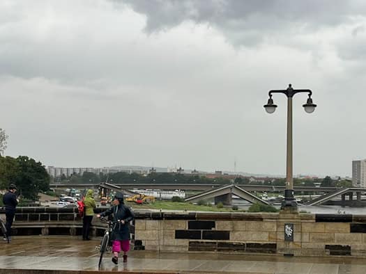 Passanten bei Regen mit Blick auf die eingestürzte Carolabrücke in Dresden. Eine Person schiebt ein Fahrrad über nasses Pflaster vor düsterem Himmel. Im Flussbett der Elbe liegen die abgebrochenen Betonteile der Brücke.