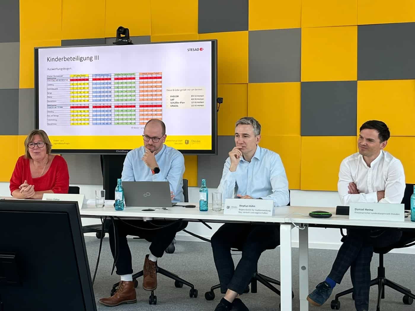 Pressekonferenz zum Carolabrücke-Neubau vor gelb-grauer Wand: Anke Lietzmann, Steven Eichler, Baubürgermeister Stephan Kühn und Stadtsprecher Daniel Heine am Tisch (v.l.). Ein Monitor im Hintergrund zeigt die Ergebnisse der Kinderbeteiligung.