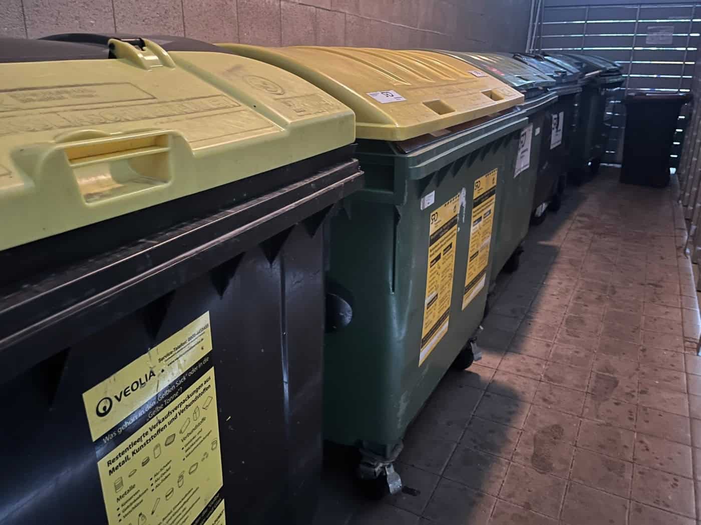 Mehrere große Mülltonnen mit gelben und dunklen Deckeln stehen aufgereiht in einem überdachten Müllraum. Auf einem Behälter ist ein Aufkleber für Wertstoffe zu sehen.