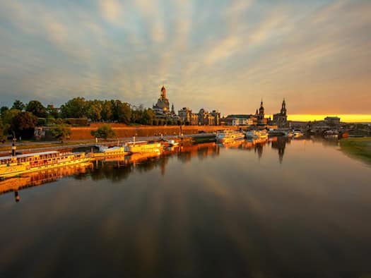 Panoramablick auf die Dresdner Altstadt bei warmem Abendlicht: Die Elbe spiegelt die historische Kulisse mit der Frauenkirche und Raddampfern am Ufer wider. Der Himmel erstrahlt in sanften Orange- und Gelbtönen mit leichten Wolkenstreifen.