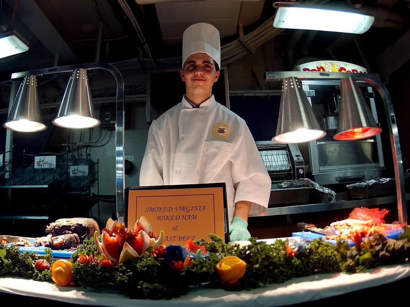 Ein junger Koch in weißer Kochjacke und hoher Kochmütze steht lächelnd hinter einer Buffet-Theke mit Fleischgerichten und kunstvoll geschnitzten Gemüsedekorationen unter Wärmelampen.