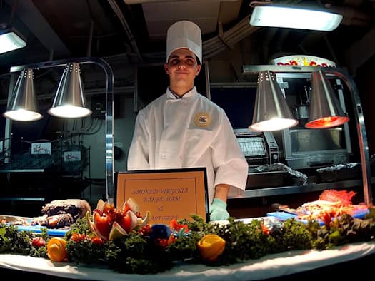 Ein junger Koch in weißer Kochjacke und hoher Kochmütze steht lächelnd hinter einer Buffet-Theke mit Fleischgerichten und kunstvoll geschnitzten Gemüsedekorationen unter Wärmelampen.