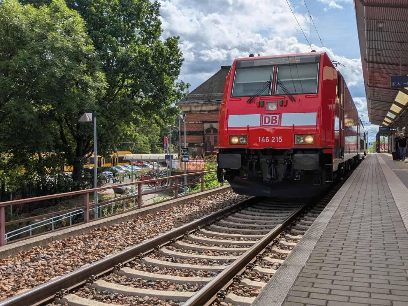Eine rote Lokomotive der Deutschen Bahn fährt am überdachten Bahnsteig des Bahnhofs Dresden-Niedersedlitz ein. Rechts warten Fahrgäste auf dem Bahnsteig, links im Hintergrund stehen Bäume und Parkplätze.