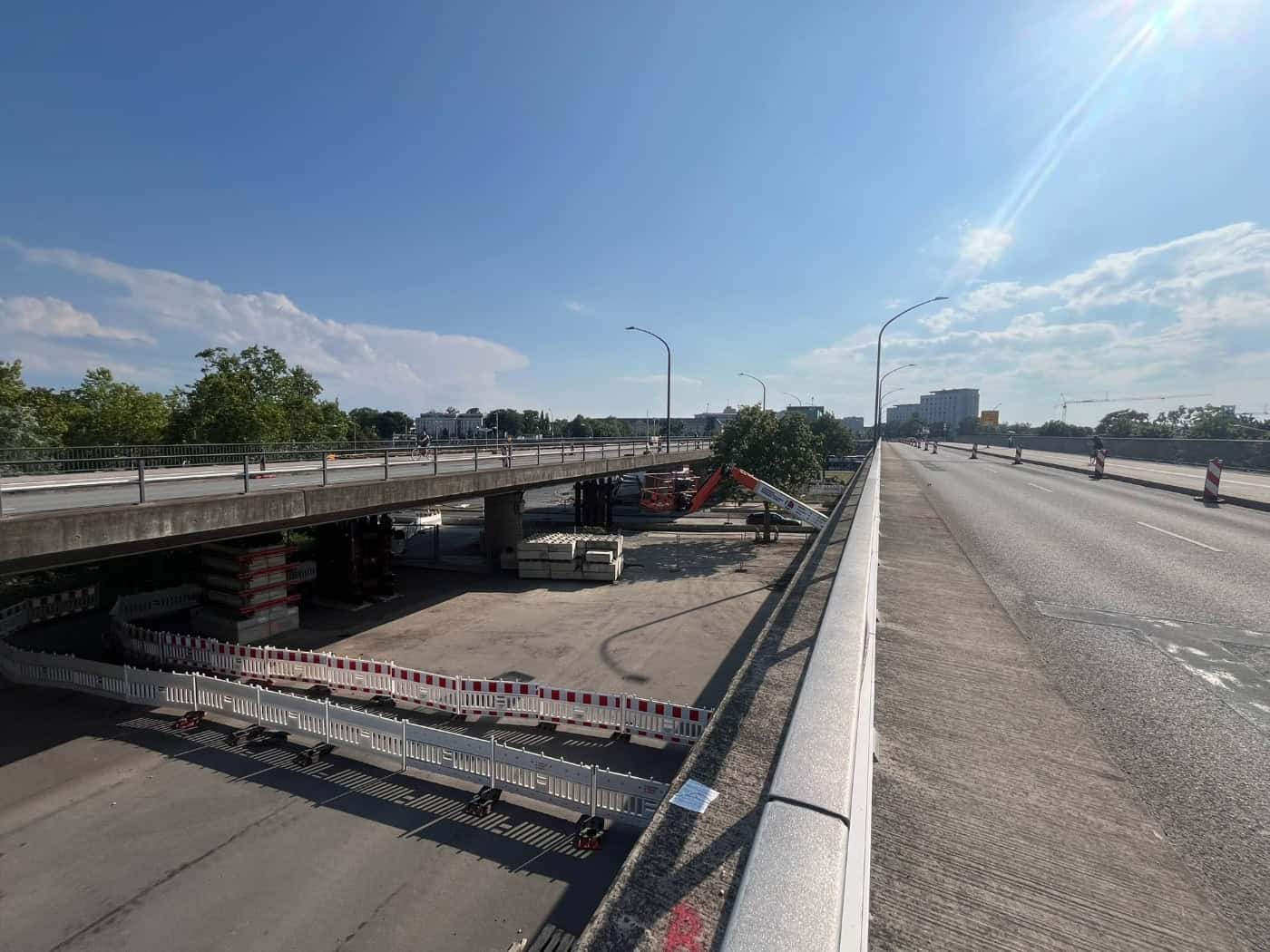 Blick entlang der Brücke Budapester Straße in Dresden bei Sonnenschein. Unter dem Betonbauwerk stehen gestapelte Notstützen und rot-weiße Absperrgitter. Die Fahrbahn oben ist weitgehend frei.