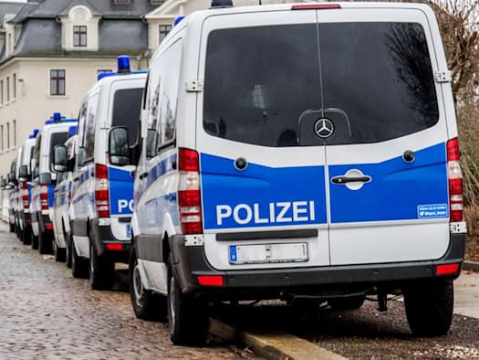 Mehrere blau-weiße Einsatzfahrzeuge der Polizei stehen hintereinander geparkt an einer Kopfsteinpflasterstraße vor einem mehrstöckigen Gebäude unter bewölktem Himmel.