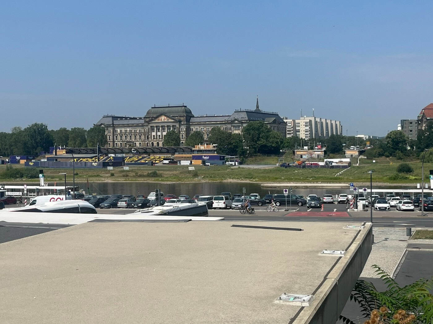 Blick von einer betonierten Plattform über einen Parkplatz am Terrassenufer und die Elbe in Dresden. Am gegenüberliegenden Ufer liegt das historische Gebäude der Staatskanzlei unter klarem blauem Himmel.
