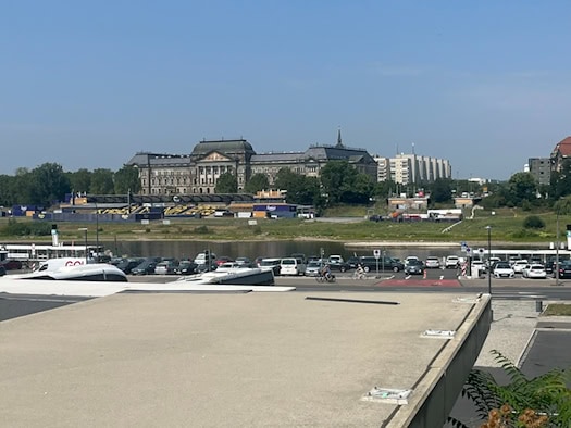 Blick von einer betonierten Plattform über einen Parkplatz am Terrassenufer und die Elbe in Dresden. Am gegenüberliegenden Ufer liegt das historische Gebäude der Staatskanzlei unter klarem blauem Himmel.