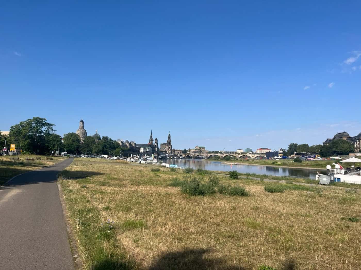 Weiter Blick über die Dresdner Elbwiesen auf die ruhige Elbe. Im Hintergrund ist bei klarem blauen Himmel die historische Altstadt mit Frauenkirche und Hofkirche zu sehen. Links im Vordergrund verläuft ein asphaltierter Fuß- und Radweg.