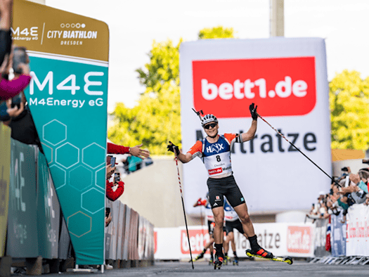 Biathlet Justus Strelow jubelt mit erhobenen Stöcken und ausgebreiteten Armen auf Skirollern beim Zieldurchlauf im Heinz-Steyer-Stadion in Dresden, umgeben von Werbebanden und applaudierenden Zuschauern.