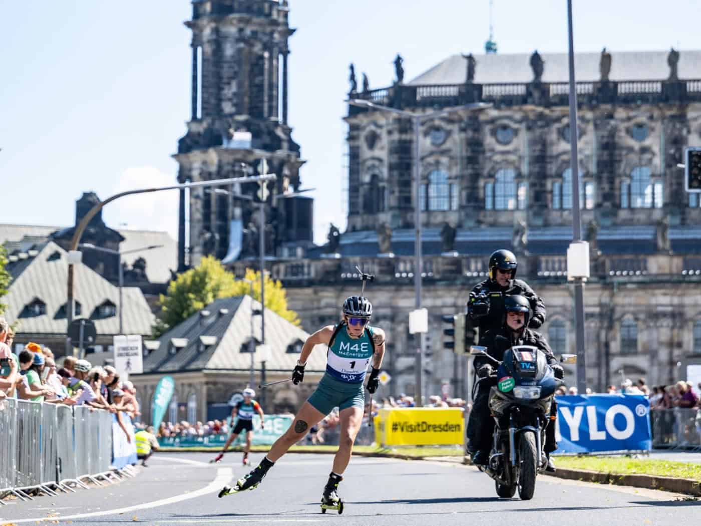 Eine Biathletin auf Skirollern fährt in Dresden während des City-Biathlons auf abgesperrter Straße. Neben ihr fährt ein Begleitmotorrad. Im Hintergrund ragen der Hausmannsturm und die Katholische Hofkirche unter blauem Himmel empor.
