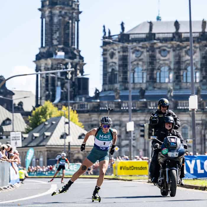 A biathlete on roller skis is racing on a closed-off street in Dresden during the City Biathlon. An escort motorcycle is riding alongside her. In the background, the Hausmannsturm and the Catholic Court Church rise against a blue sky.