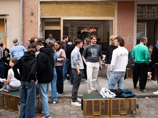 Viele junge Menschen stehen und sitzen vor der Galerie goldene Pforte auf dem Gehweg der Martin-Luther-Straße. Im Schaufenster leuchtet ein Neonschriftzug mit dem Galerie-Namen.