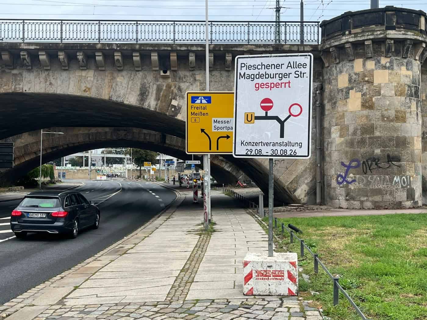 Ein weißes Hinweisschild warnt vor der Sperrung der Pieschener Allee und Magdeburger Straße wegen einer Konzertveranstaltung vom 29. bis 30.08.26. Im Hintergrund führt eine Straße unter einer alten Steinbrücke hindurch.