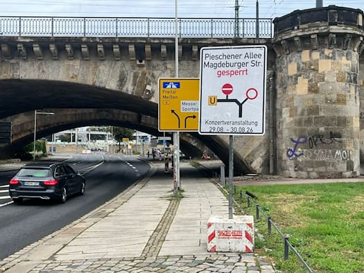 Ein weißes Hinweisschild warnt vor der Sperrung der Pieschener Allee und Magdeburger Straße wegen einer Konzertveranstaltung vom 29. bis 30.08.26. Im Hintergrund führt eine Straße unter einer alten Steinbrücke hindurch.