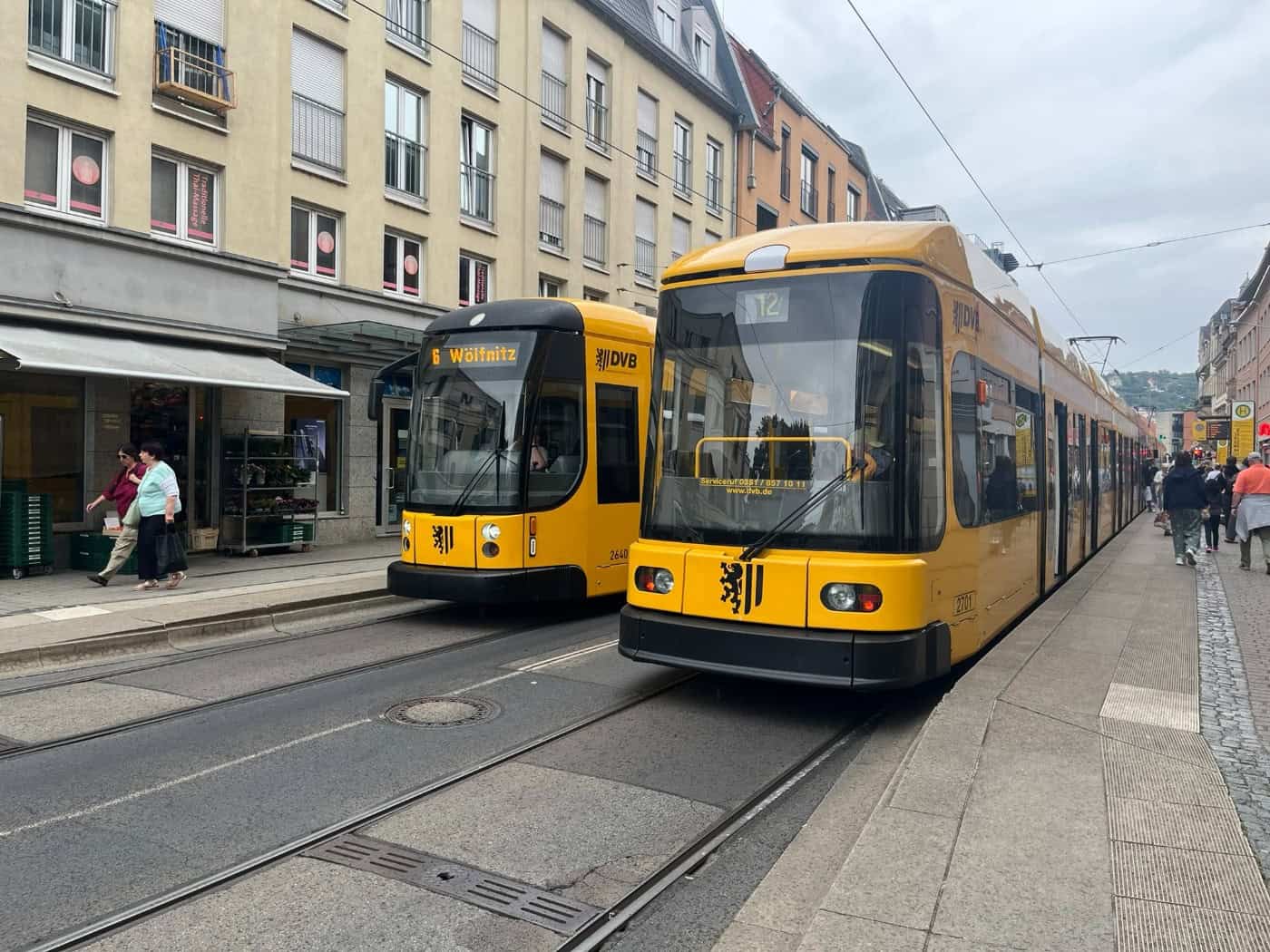 Zwei gelbe Straßenbahnen der Dresdner Verkehrsbetriebe (DVB) auf den Schienen in einer belebten Dresdner Einkaufsstraße. Vorne rechts ist die Linie 12 zu sehen, links daneben die Linie 6 mit Ziel Wölfnitz.