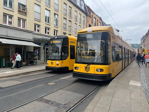 Zwei gelbe Straßenbahnen der Dresdner Verkehrsbetriebe (DVB) auf den Schienen in einer belebten Dresdner Einkaufsstraße. Vorne rechts ist die Linie 12 zu sehen, links daneben die Linie 6 mit Ziel Wölfnitz.
