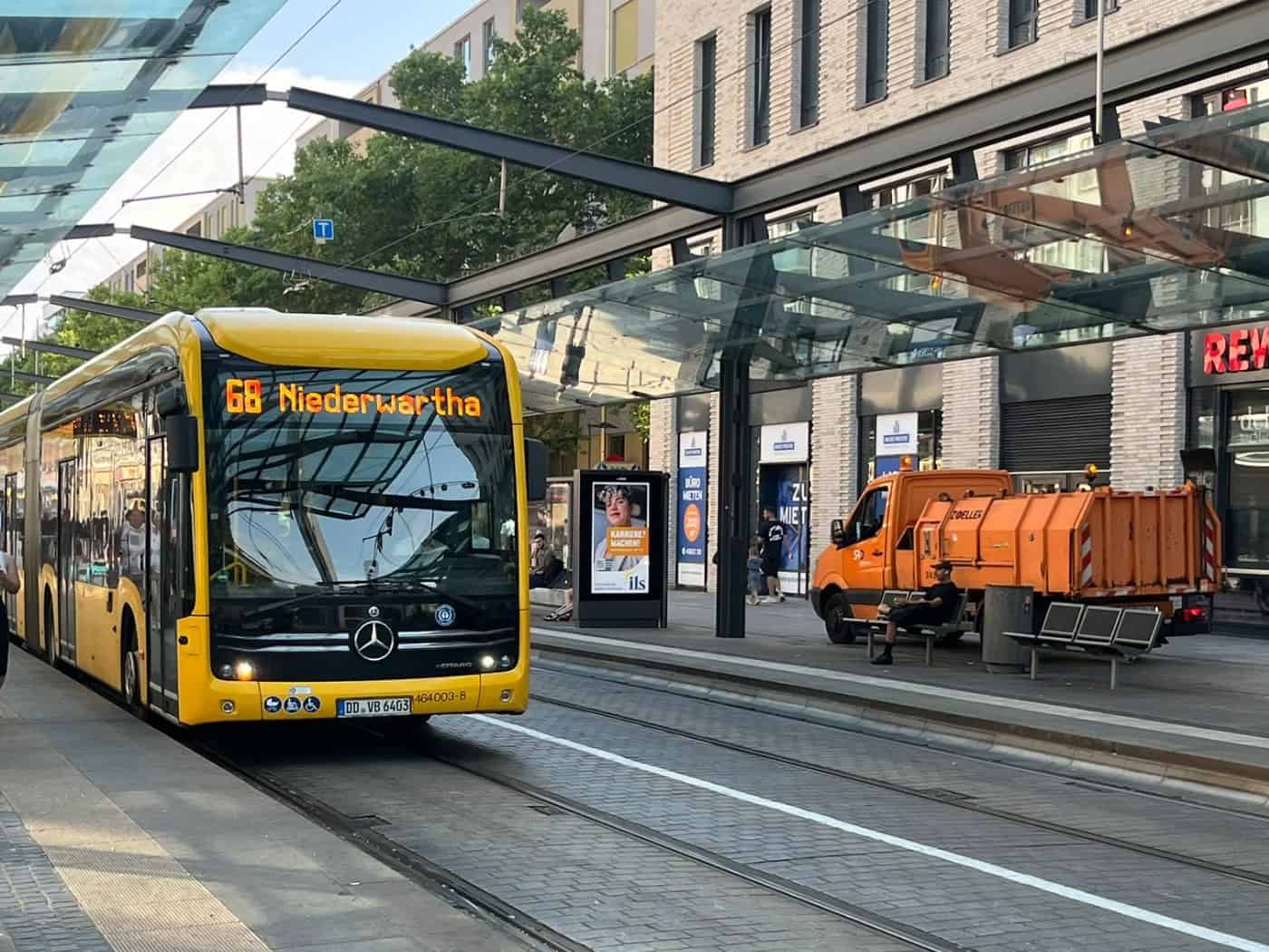 Ein gelber Linienbus der DVB mit der Anzeige „68 Niederwartha“ steht an einer überdachten Haltestelle in Dresden. Fahrgäste warten auf dem Bahnsteig, im Hintergrund ist ein Gebäude mit REWE-Filiale zu sehen.