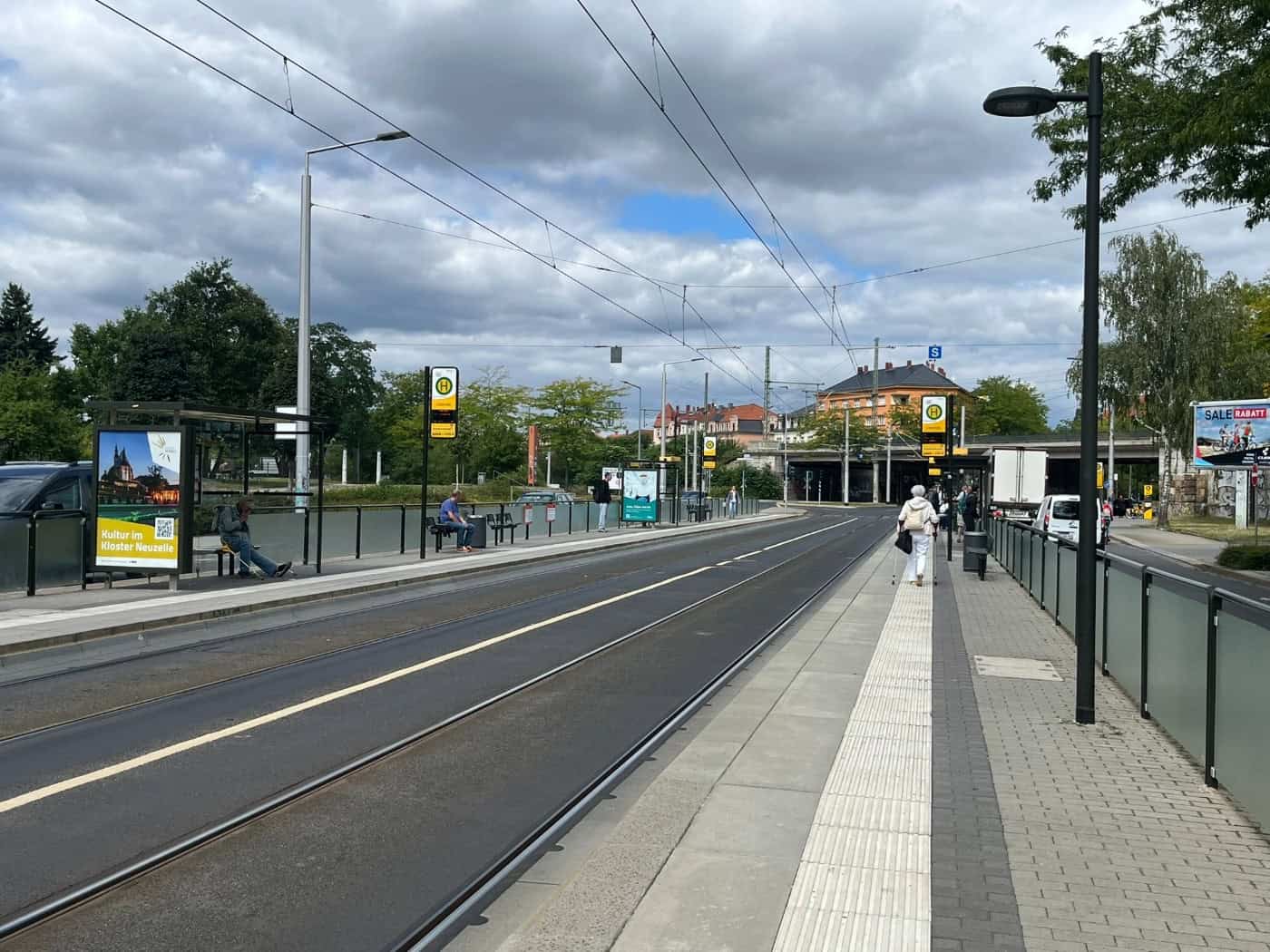Blick entlang der leeren Straßenbahngleise an der DVB-Haltestelle Liststraße in Dresden-Pieschen mit gläsernen Fahrgastunterständen, S-Bahn-Unterführung und Wolkenhimmel im Hintergrund.