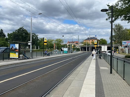 Blick entlang der leeren Straßenbahngleise an der DVB-Haltestelle Liststraße in Dresden-Pieschen mit gläsernen Fahrgastunterständen, S-Bahn-Unterführung und Wolkenhimmel im Hintergrund.