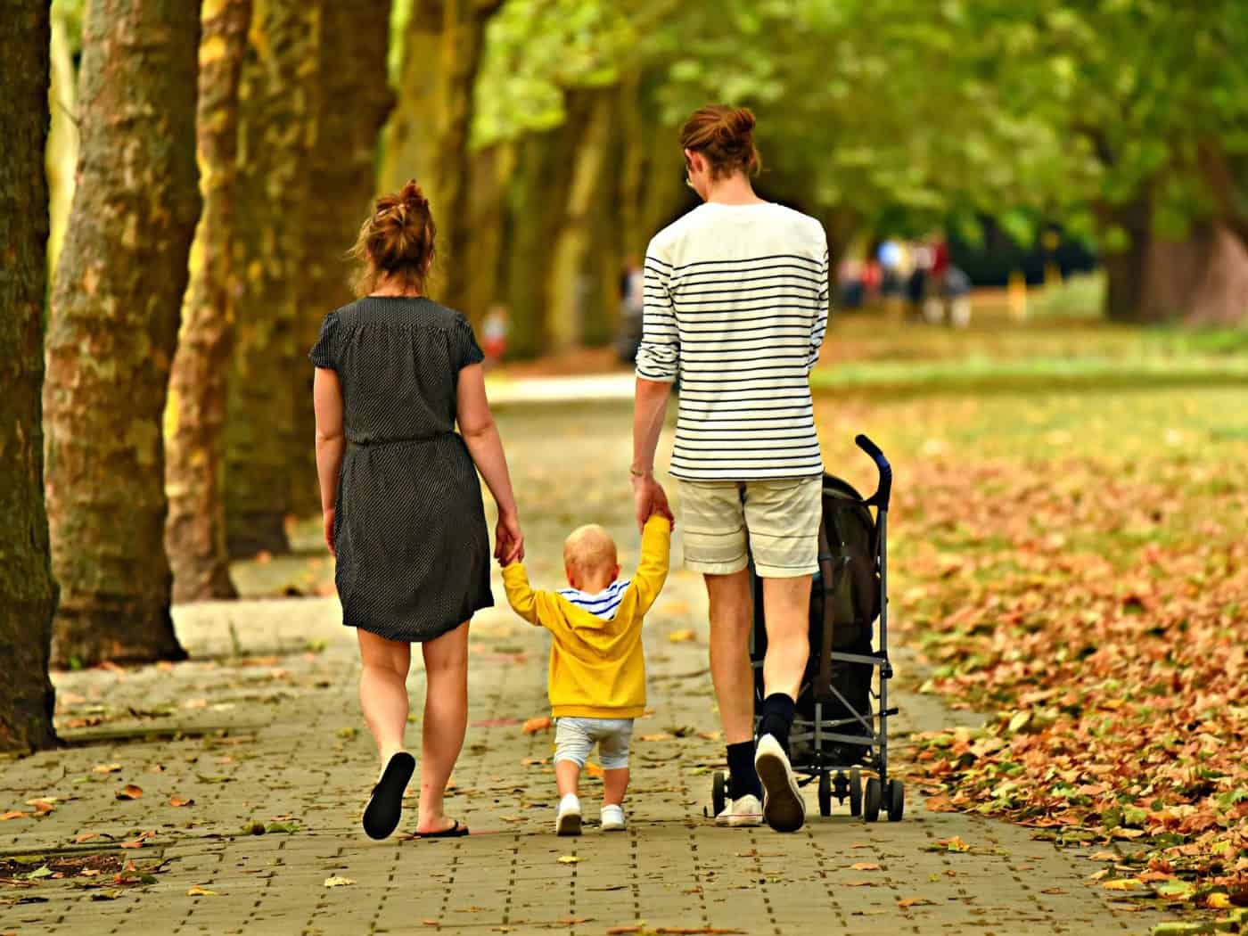 Rückansicht einer dreiköpfigen Familie, die zusammen auf einem von Bäumen gesäumten Weg spaziert: Die Eltern halten ihr kleines Kind an den Händen, während der Vater einen Kinderwagen schiebt.