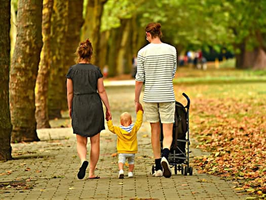 Rückansicht einer dreiköpfigen Familie, die zusammen auf einem von Bäumen gesäumten Weg spaziert: Die Eltern halten ihr kleines Kind an den Händen, während der Vater einen Kinderwagen schiebt.