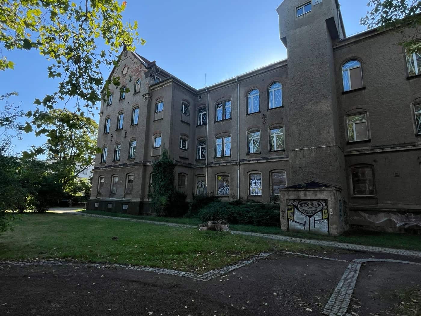 Ein mehrstöckiges, sanierungsbedürftiges historisches Gebäude mit grauem Verputz, Rundbogenfenstern und Graffiti-Verzierungen hinter einer Rasenfläche an der Löbtauer Straße in Dresden.